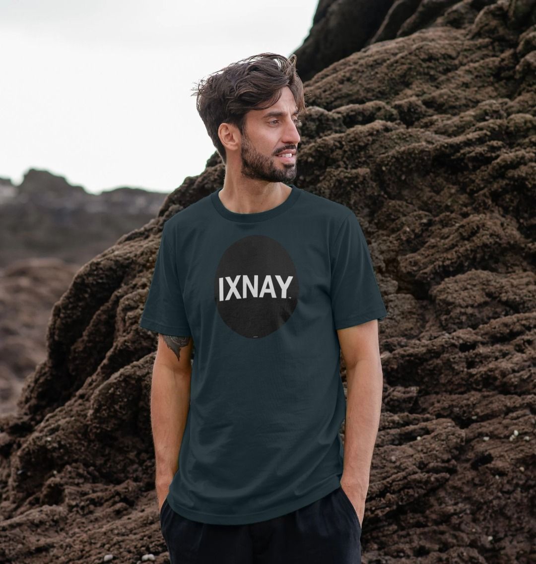 Man in front of beachside rock formation-wearing dark gray Printed Men's T-Shirt with Original Black Round IXNAY Logo to represent the artist and musician IXNAY