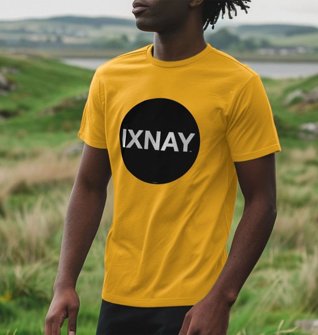 Black man standing in beautiful grassy dunes near a body of water wearing a mustard yellow Printed Men's T-Shirt with Original Black Round IXNAY Logo to represent the artist and musician IXNAY