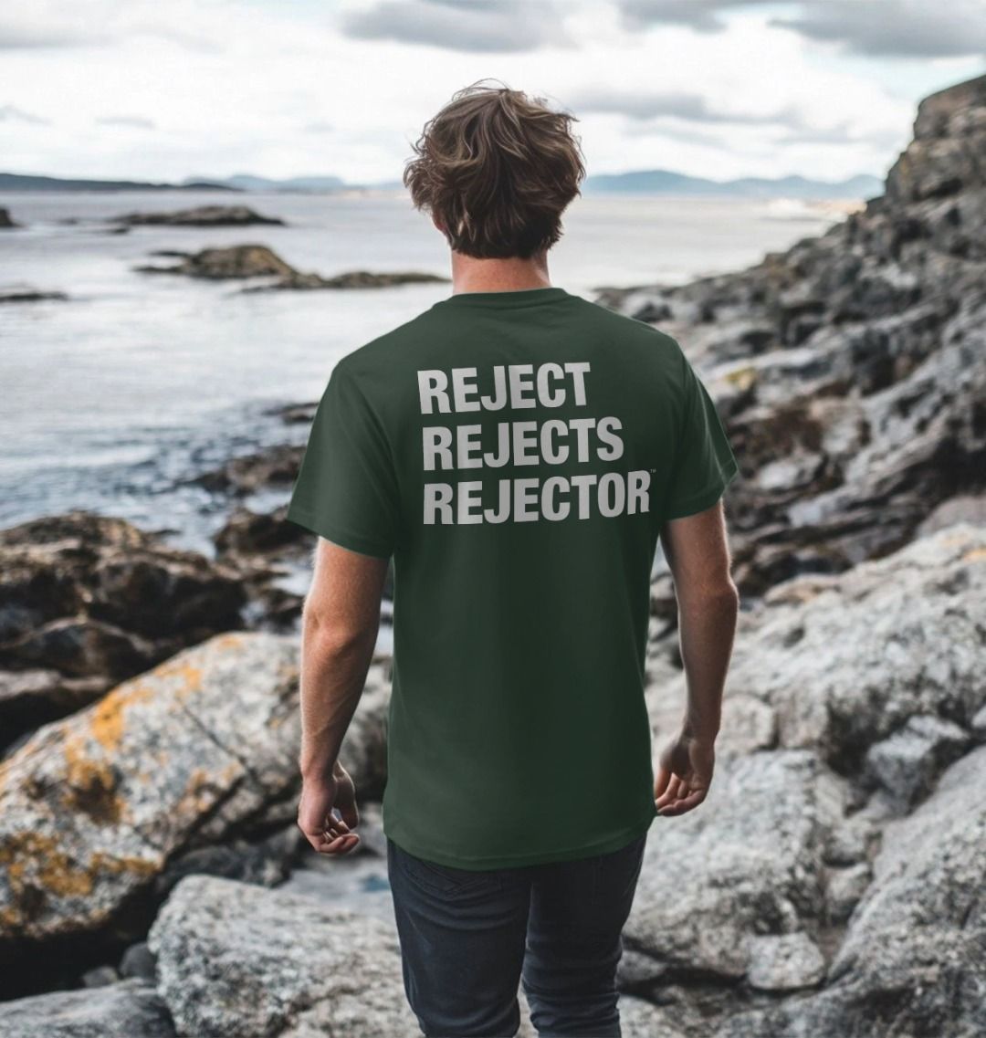 Man standing with his back to camera on a rocky beach looking out to sea wearing a dark gray Printed Men's T-Shirt with Original Black Round IXNAY Logo with the phrase REJECT REJECTS REJECTOR on back  to represent the artist and musician IXNAY