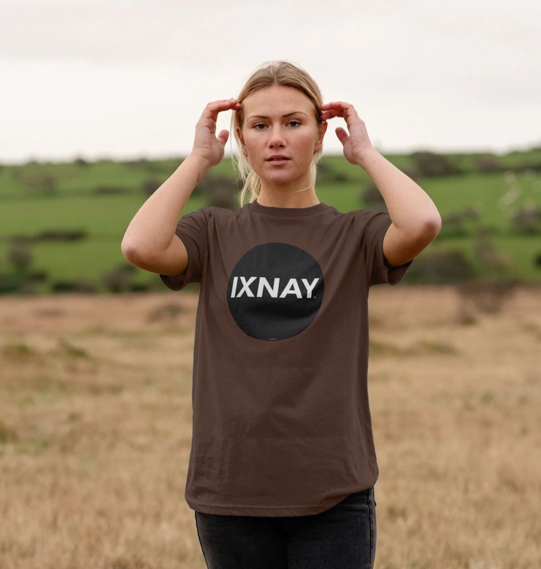 Woman in a large field wearing a chocolate brown Printed Men's T-Shirt with Original Black Round IXNAY Logo to represent the artist and musician IXNAY
