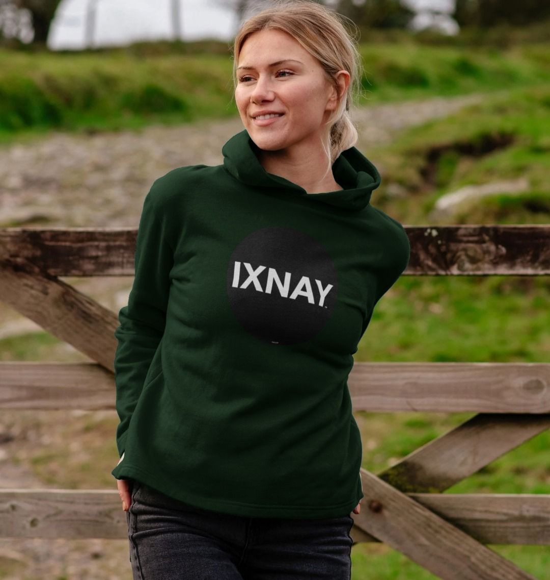 woman in front of wooden farm gate wearing womens dark green evergreen organic pullover hoodie with original black round IXNAY logo
